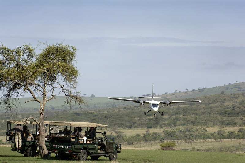 A light aircraft on a bush airstrip with an open-sided safari vehicle waiting under a tree in the foreground